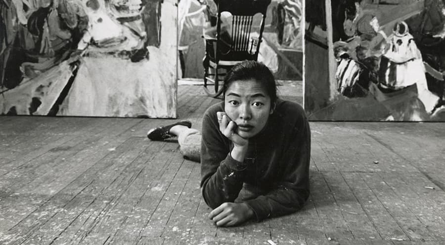 Woman lying on the floor with her hand on her chin looking at the camera with two large paintings behind her.